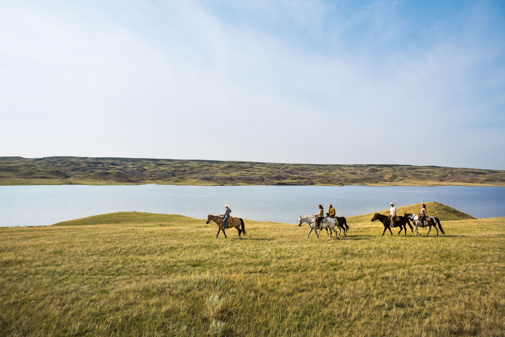 People horseback riding during the day outside on the grass by the water in Saskatchewan Canada at La Reata Ranch